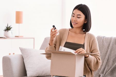 Beauty Box Concept. Portrait of smiling asian woman received delivery package, holding foundation base for makeup, unpacking cardboard parcel with cosmetics product, sitting on the couch at home