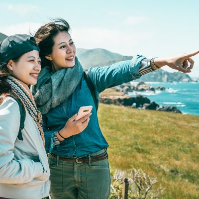 close friends looking into the ocean and one girl pointing to somewhere with a phone held by a background of stunning coast.