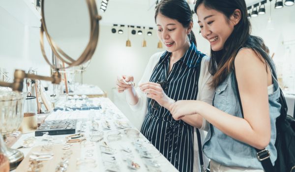 two asian women customer in busy clothes shop shopping accessories taking up from table. beautiful smiling young ladies best friends buying choosing earrings on desk in fashion modern store tokyo